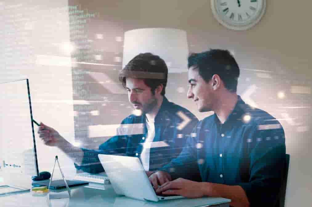 two men sitting at the table, looking at the computer screen, one in pointing on the screen with pen