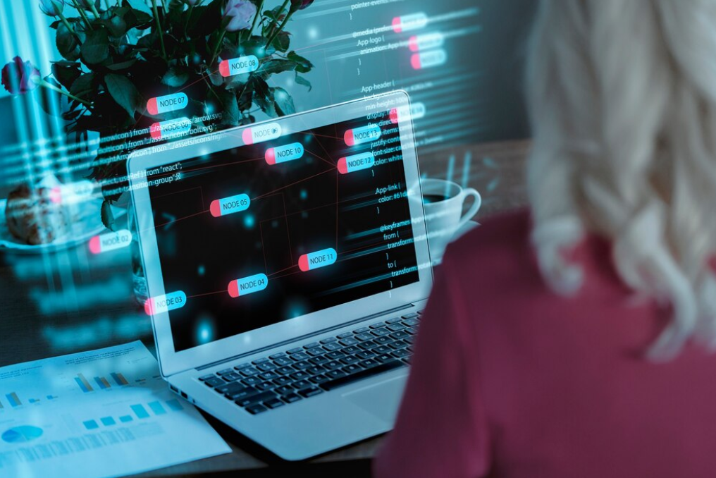 woman sits in front of the opened laptop, the sheet with graphs on the table, and code on the fond